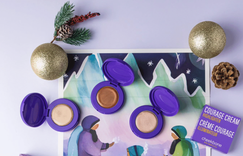 Three open cream highlighters in purple compacts arranged on a winter-themed illustration, surrounded by holiday ornaments and a pine cone.