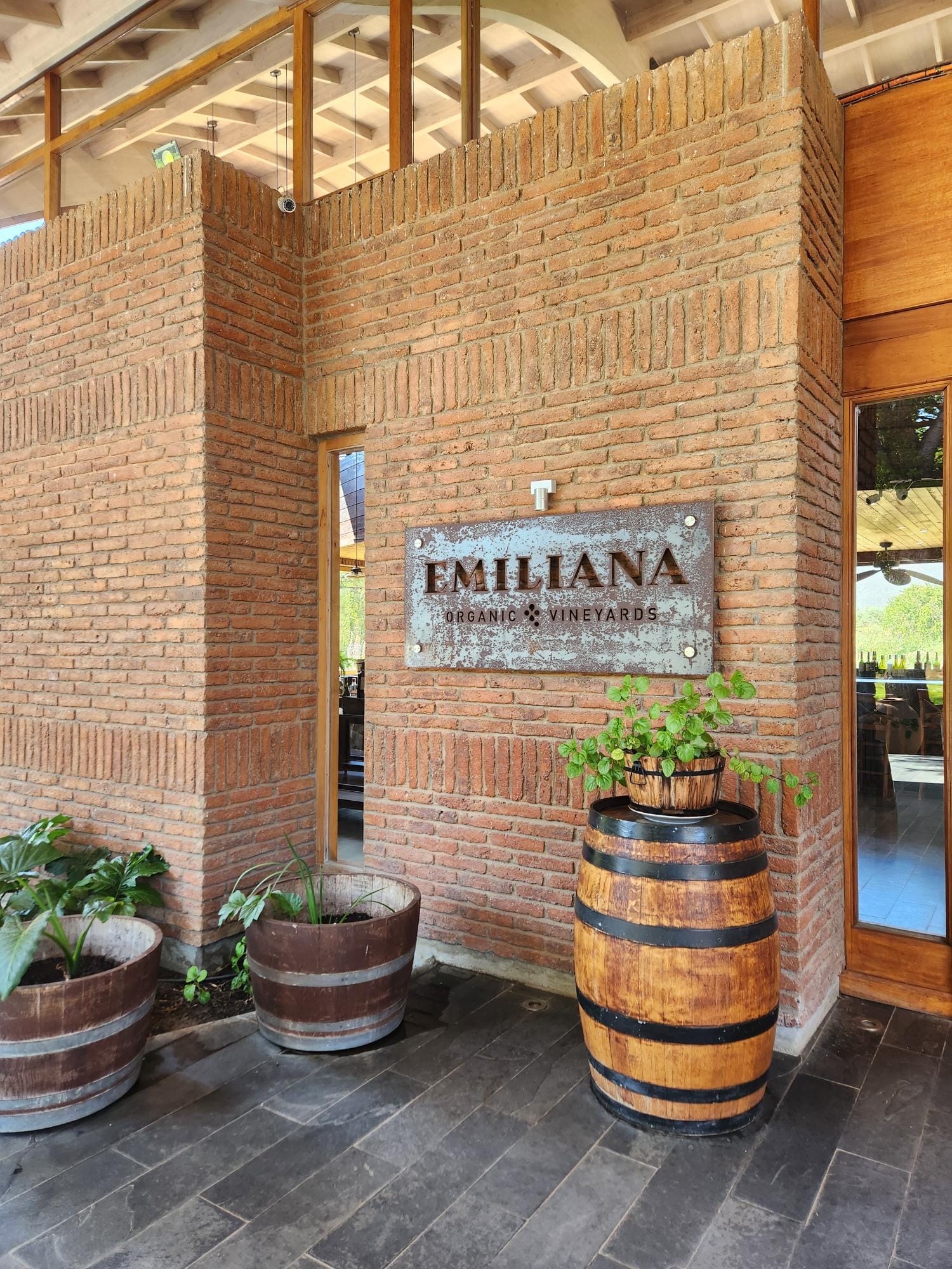 Brick entryway of Emiliana Organic Vineyards featuring a metal sign, wooden barrels used as planters, and potted plants beside a wooden door.