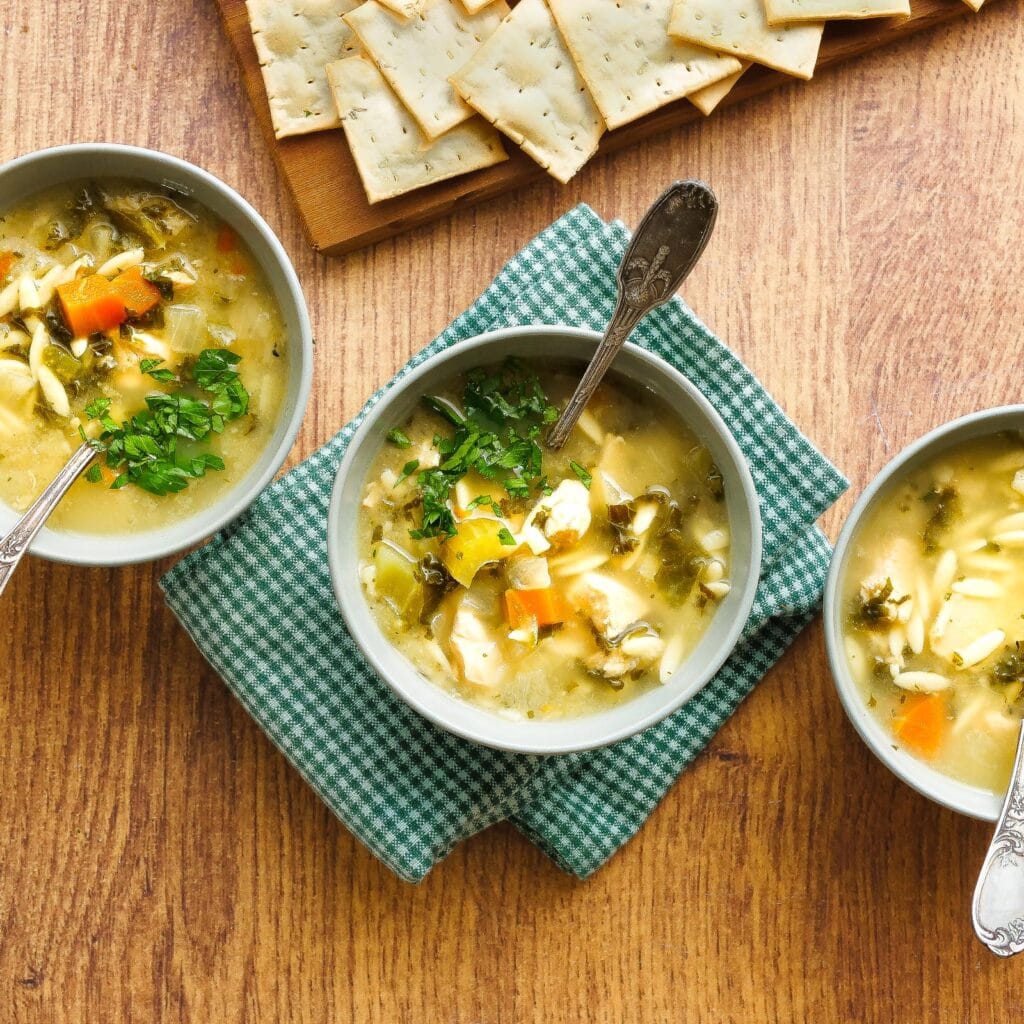 Three bowls of Goodly chicken and vegetable soup on a wooden table, each topped with fresh herbs, with saltine crackers on a board in the background.