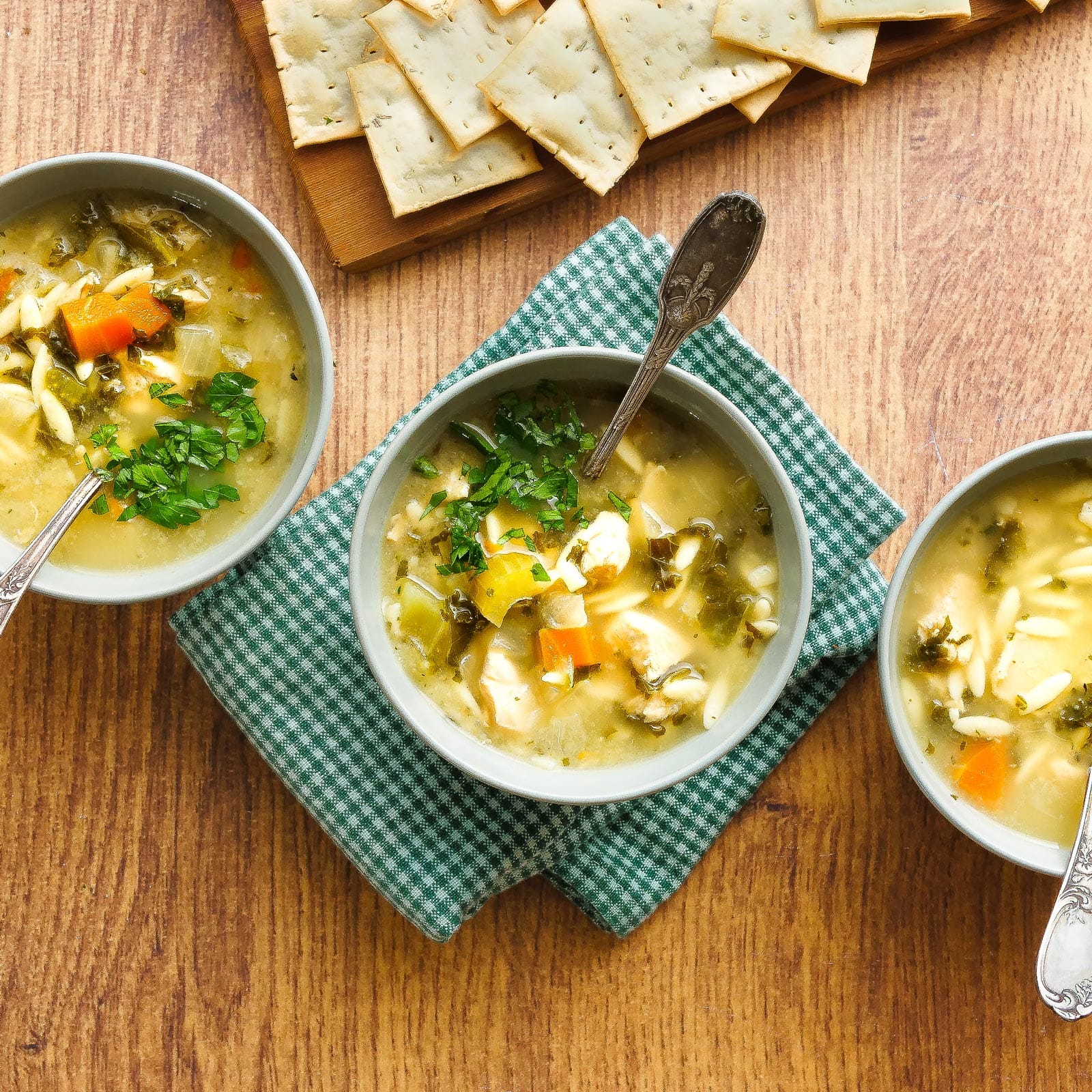 Three bowls of Goodly chicken and vegetable soup on a wooden table, each topped with fresh herbs, with saltine crackers on a board in the background.