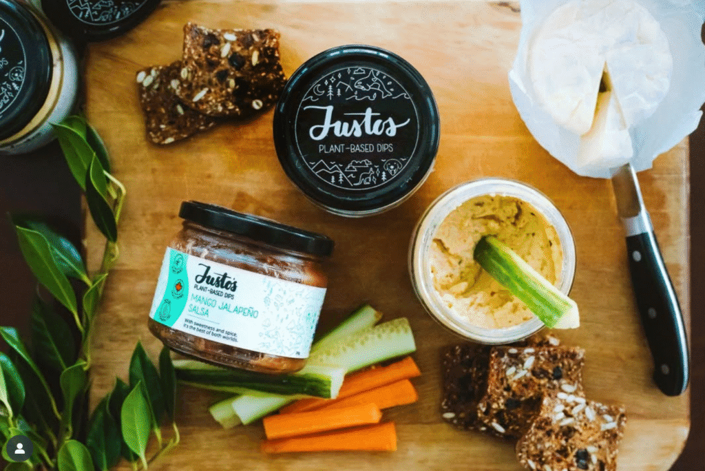 Jars of Justo’s plant-based dips displayed on a wooden board with sliced vegetables, crackers, cheese, and fresh greenery.