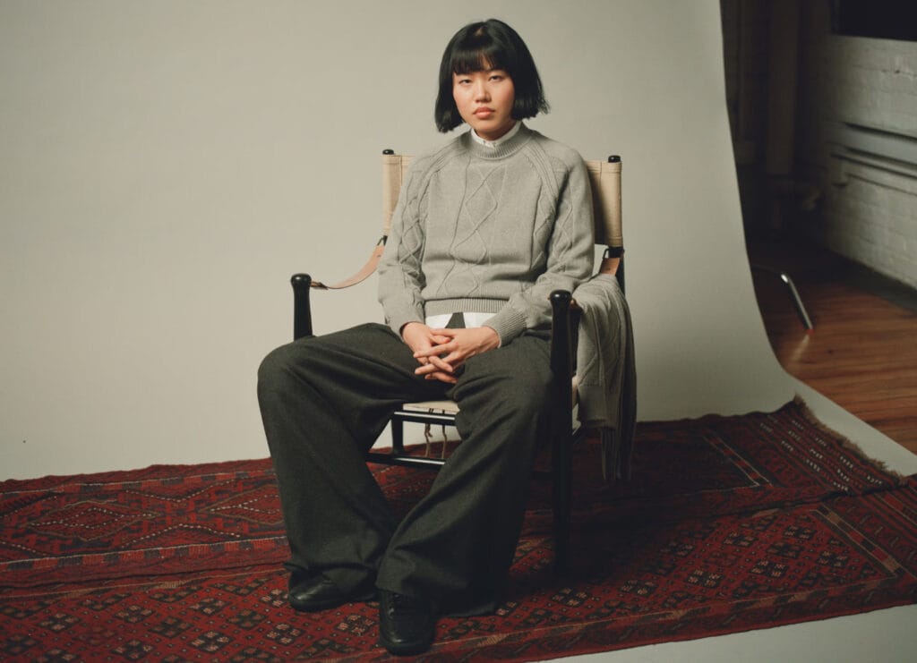 A person with a short bob haircut sits on a wooden chair atop a patterned red rug, wearing a grey cable-knit sweater, a white shirt, and loose dark trousers, posed against a simple studio backdrop.