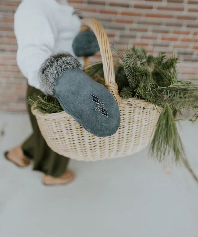 A person wearing blue suede Manitobah mittens with fur trim while holding a wicker basket filled with pine branches.