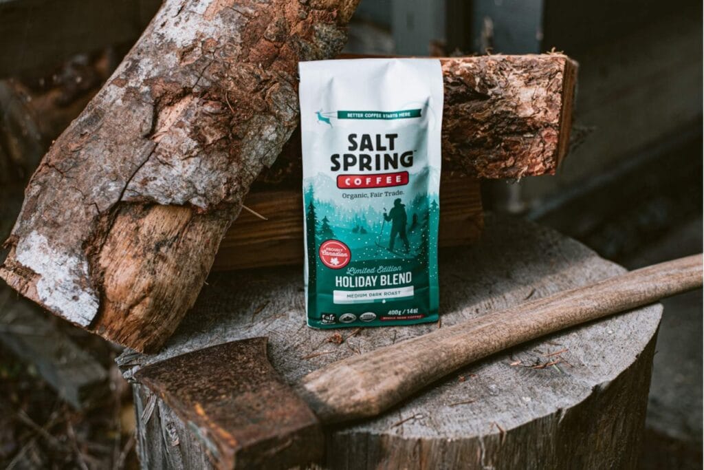 A bag of Salt Spring Coffee Holiday Blend resting on a tree stump beside stacked firewood and an axe handle.