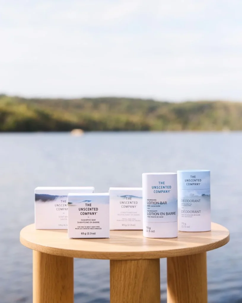 A collection of The Unscented Company soap bars, lotion bar, conditioner bar, and deodorant displayed on a small wooden table by a lakeside.